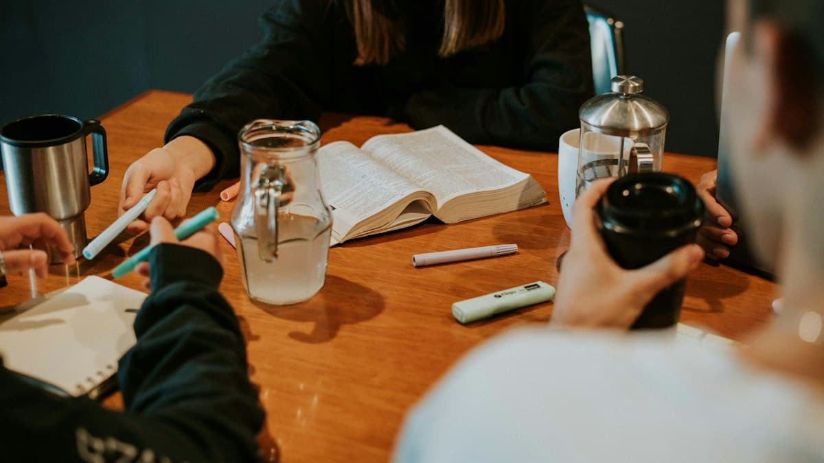 Small group sitting around a wooden table with an open Bible, highlighters and coffee cups; only hands are in focus