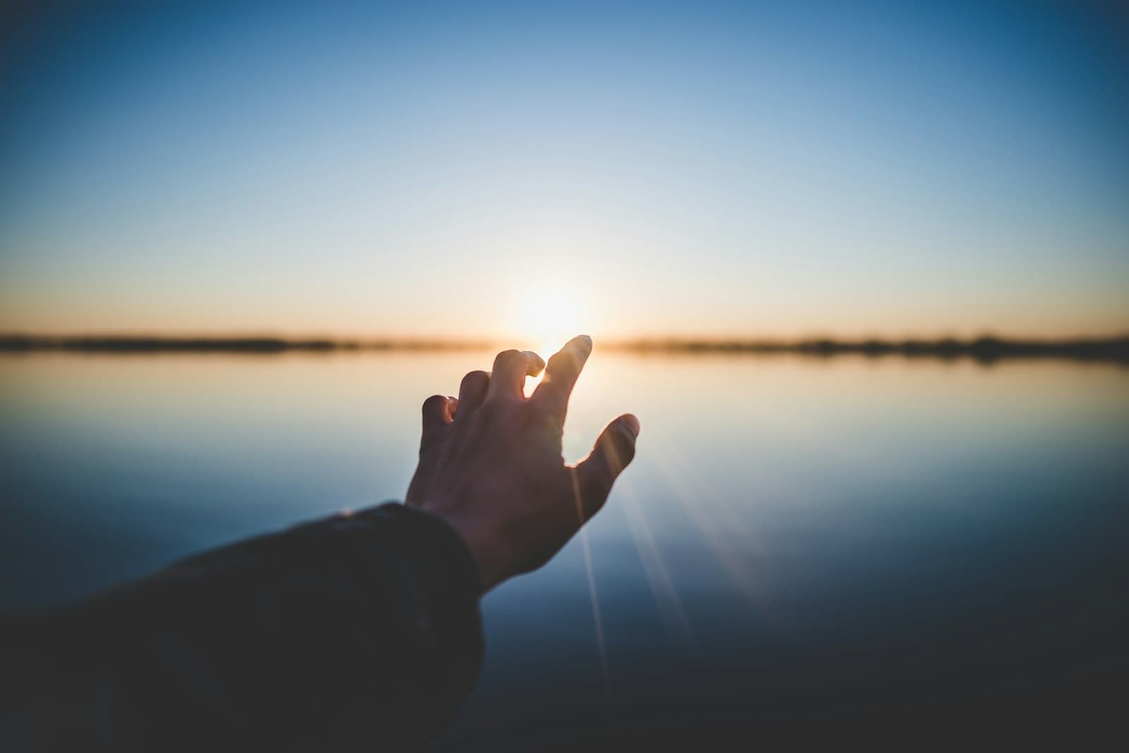 A hand reaching toward the low sun over still water