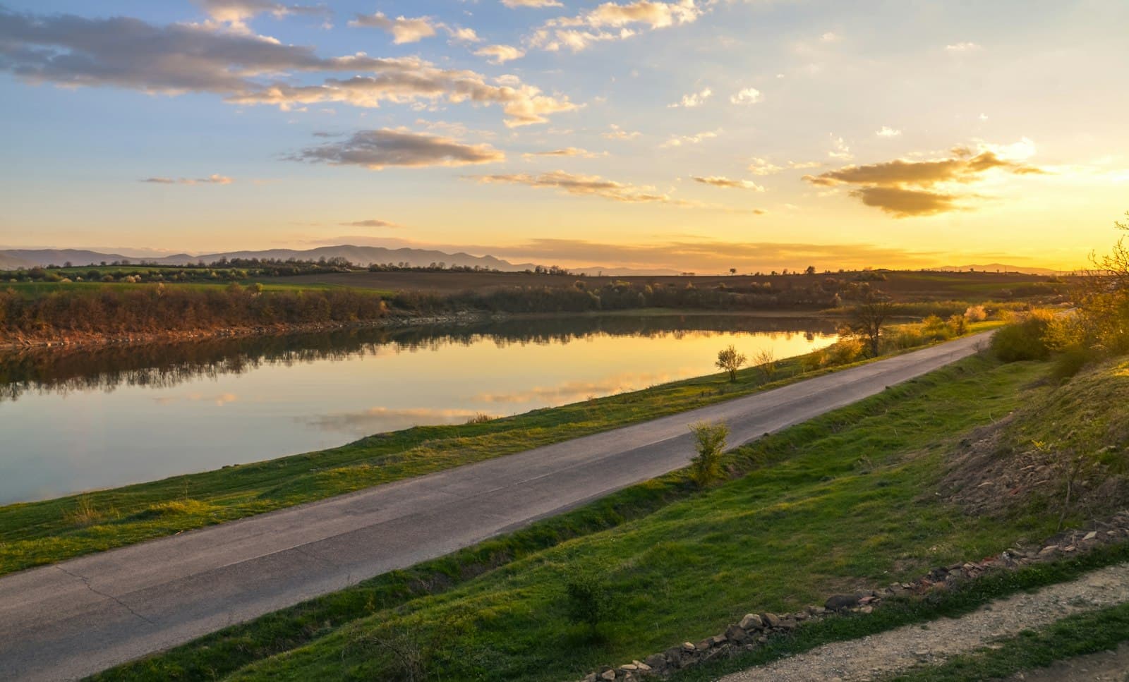 A road beside a calm river at sunset, with golden clouds over green fields