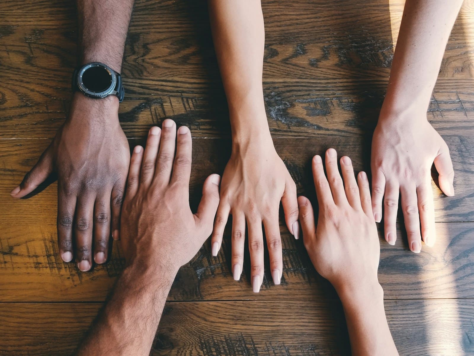 Five hands of different people resting side by side on a wooden table
