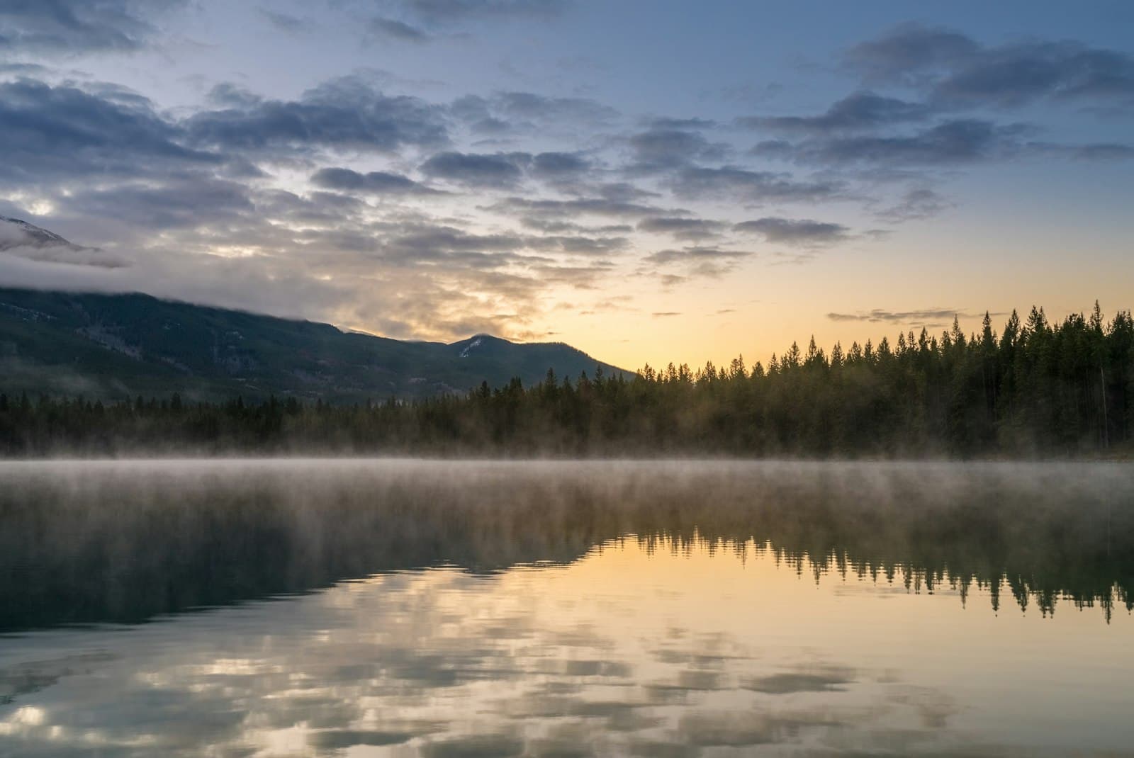Still lake at sunrise reflecting clouds, with mist over the water and a forested shore