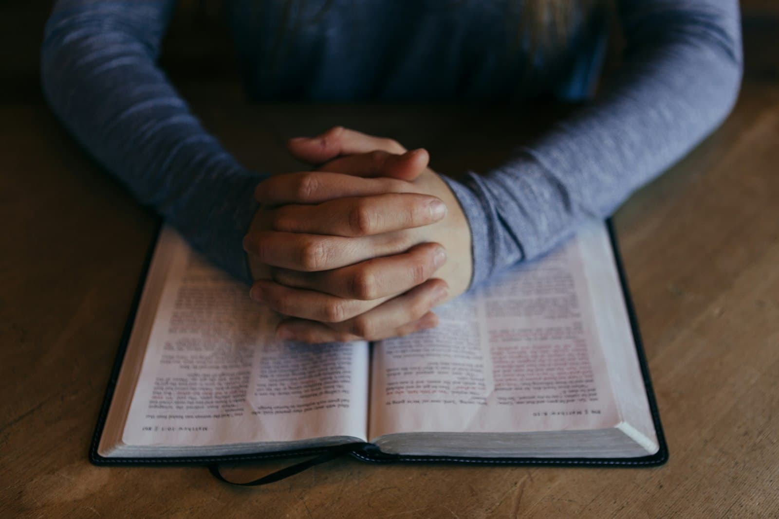 Folded hands resting on an open Bible on a wooden table