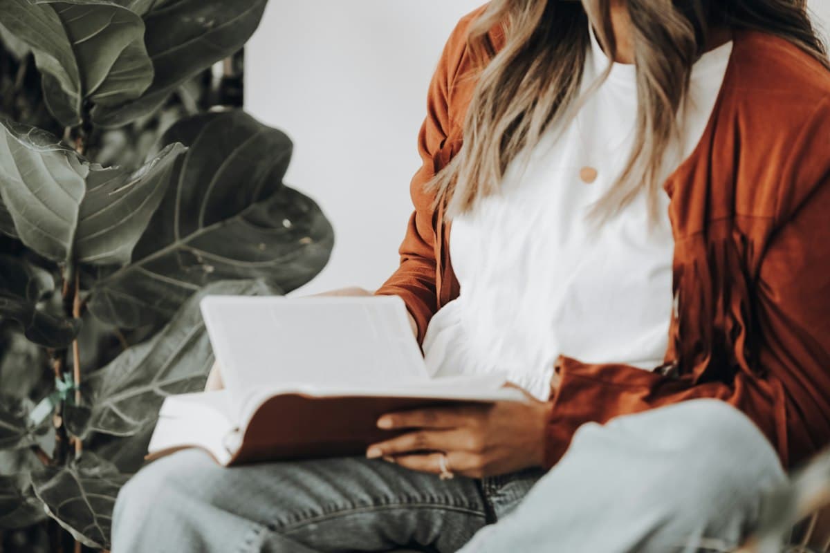 Woman sitting beside a large leafy plant, reading an open book on her lap